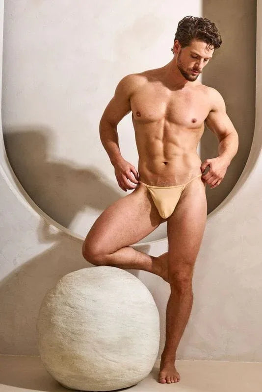 Athletic man modeling beige men's thong underwear in a modern, minimal studio setting.