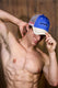 Shirtless fit male model in blue Coyote logo trucker hat posing indoors, wood background.
