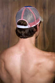 Shirtless man from behind wearing a mesh red and white trucker hat against wood background