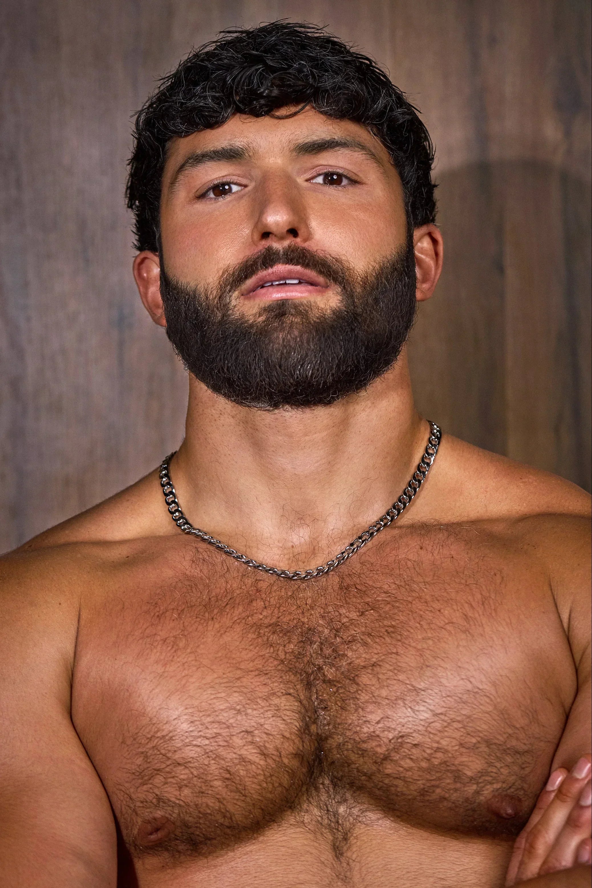 Muscular, bearded man with chest hair wearing a chain necklace against a wood background.