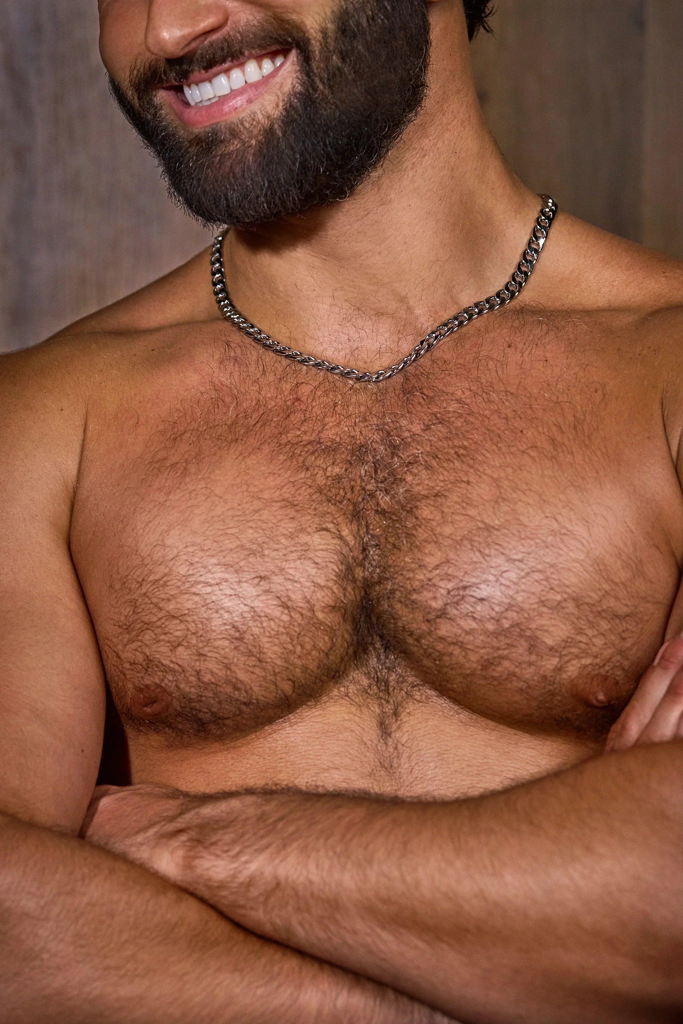 Shirtless bearded man with hairy chest and chain necklace, arms crossed, smiling