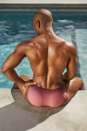 Muscular man in pink swim briefs sitting poolside, highlighting men's swimwear fashion