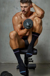 Athletic man in black underwear and Coyote socks lifting a dumbbell on a gym bench.