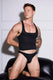 Muscular man in black tank top and low-rise briefs, showcasing athletic build and tattoos on arm and legs.