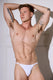 Man in white thong underwear, showcasing fit physique and arm tattoos against white paneling