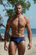 Muscular man in blue swim briefs with gold stripes, standing on a sunny beach