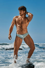 Muscular man in light blue swim brief standing in ocean surf on a sunny beach