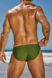 Man in green swim brief at the beach, showing athletic back and arm tattoo