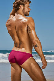 Man wearing magenta swim briefs posing on beach, showing back and arm tattoos under sunlight