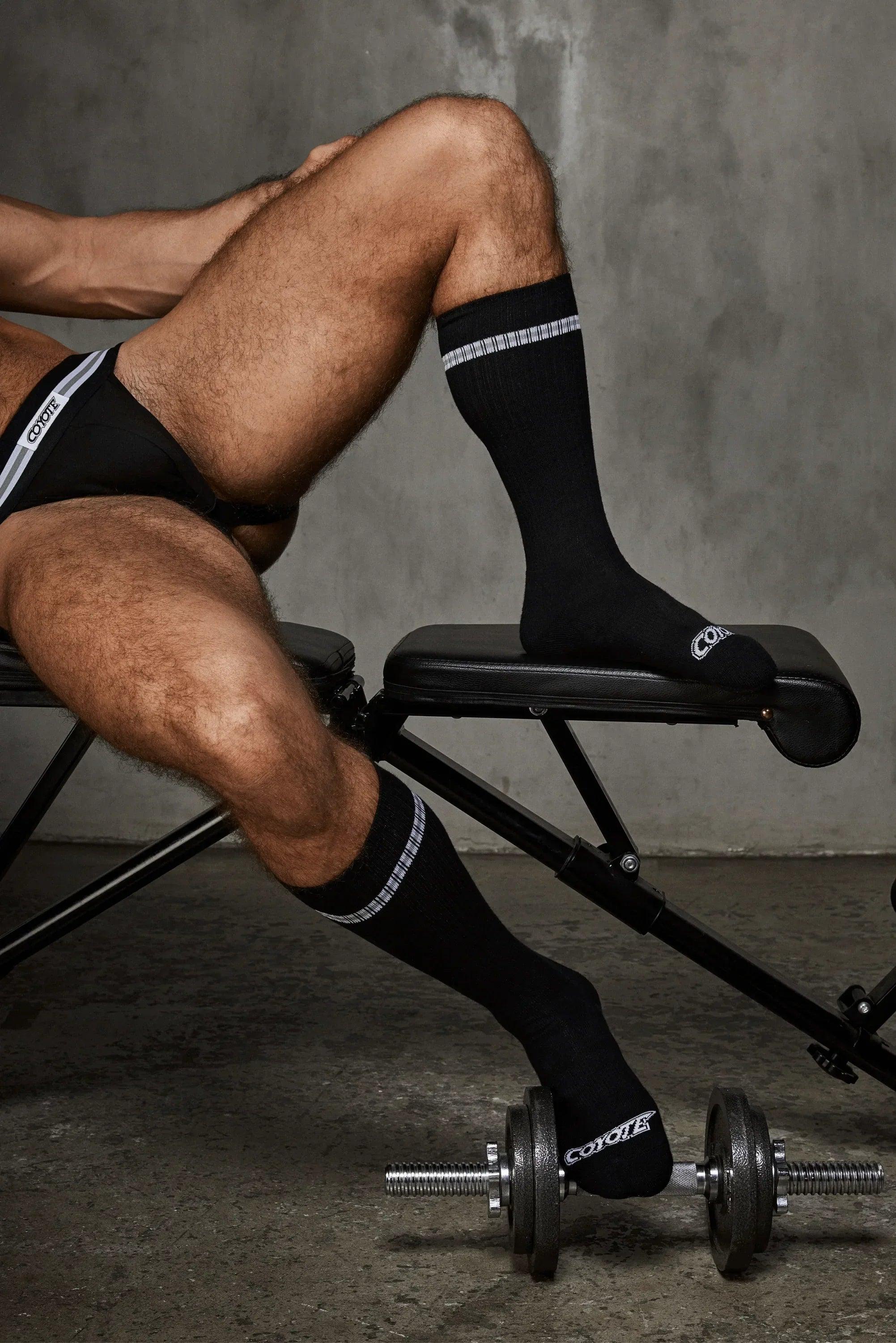 Man in black jockstrap and black athletic socks sitting on workout bench, dumbbell on floor