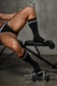 Man in black jockstrap and black athletic socks sitting on workout bench, dumbbell on floor
