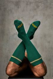Man wearing ribbed green knee-high socks with yellow logo and striped trim, legs crossed.
