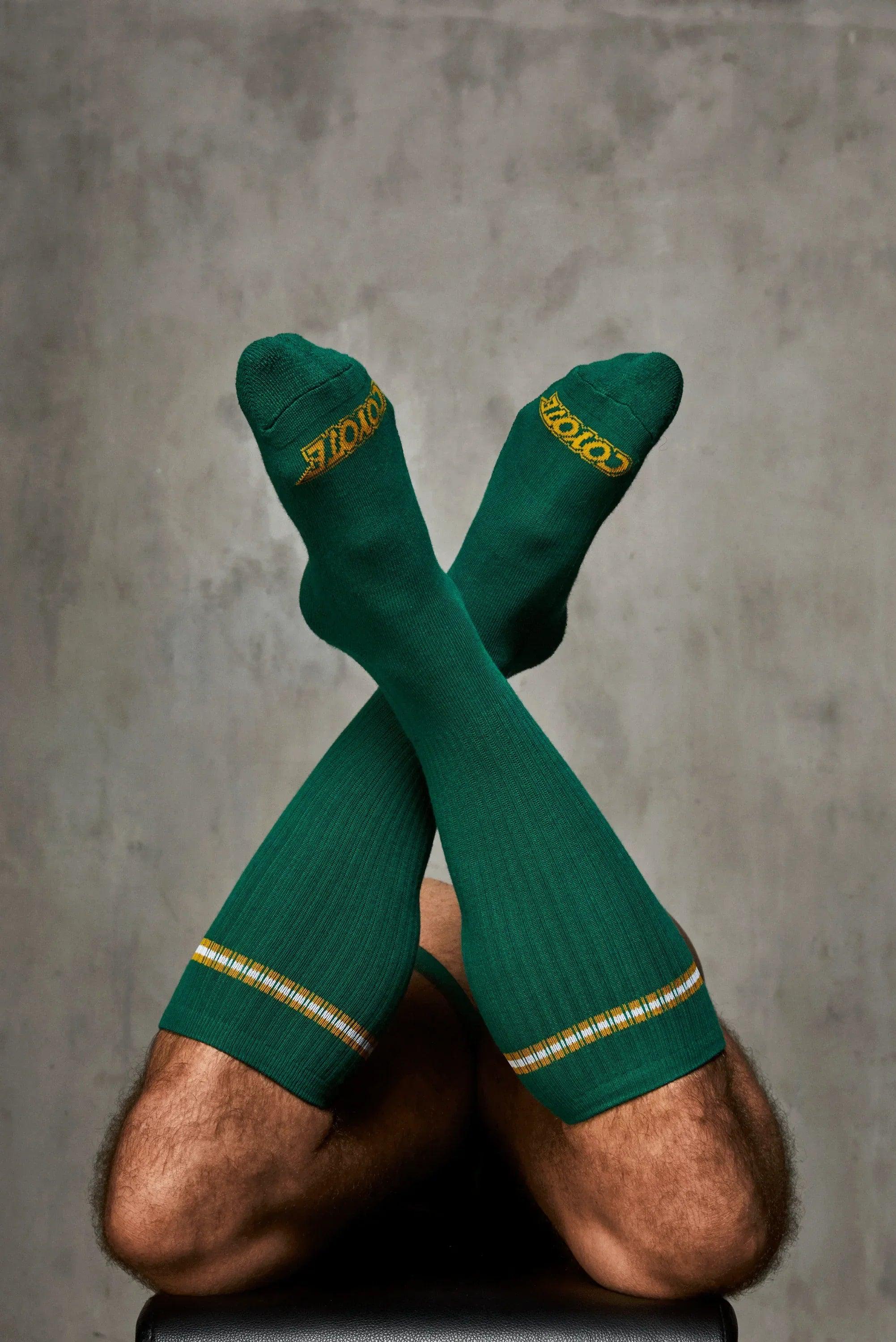 Man wearing ribbed green knee-high socks with yellow logo and striped trim, legs crossed.
