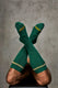 Man wearing ribbed green knee-high socks with yellow logo and striped trim, legs crossed.