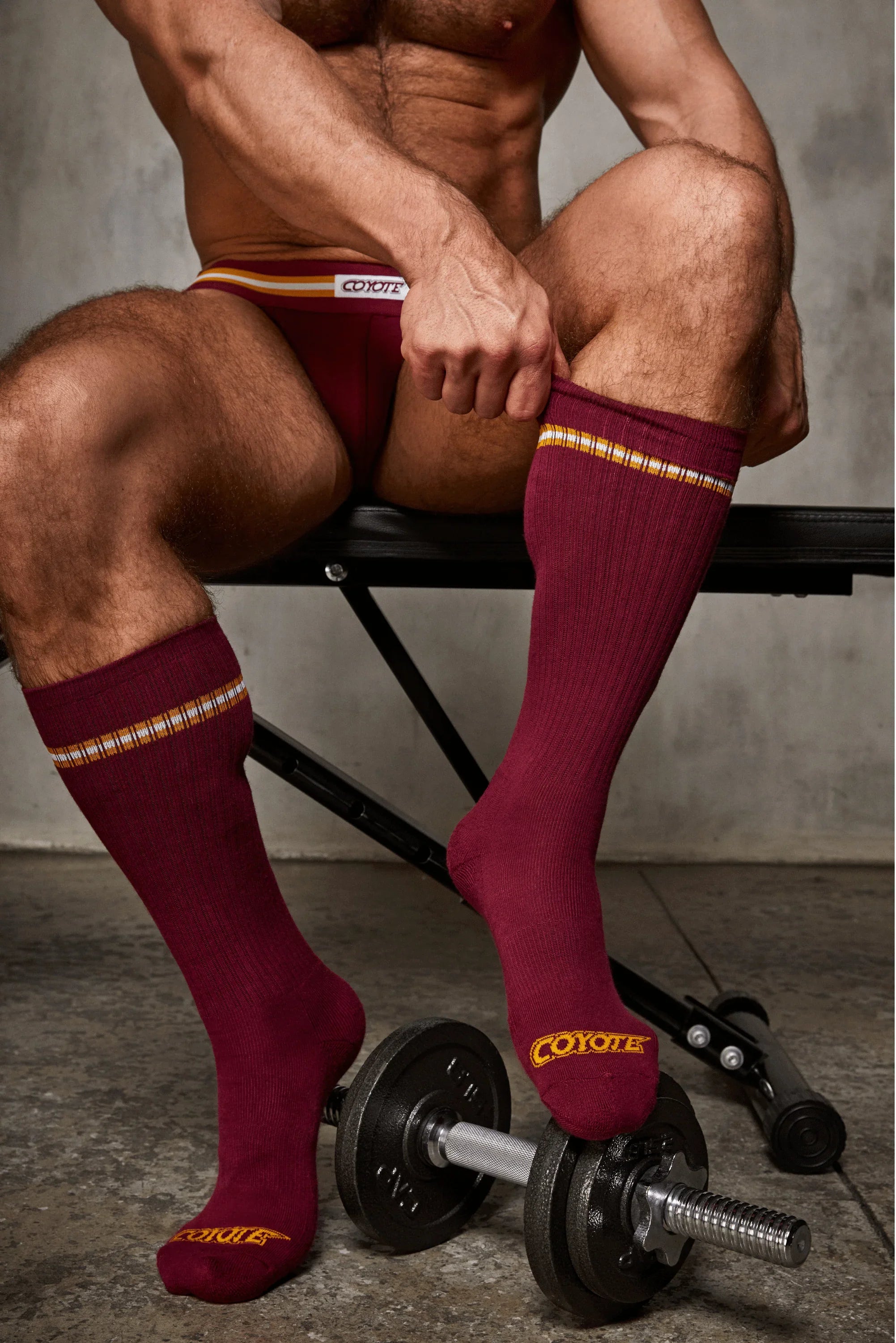 Man wearing maroon athletic underwear and socks, seated on gym bench with dumbbell